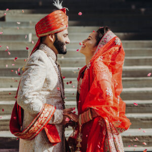 Indian groom dressed in white Sherwani and red hat with stunning bride in red lehenga stand and hold each hands walking outside